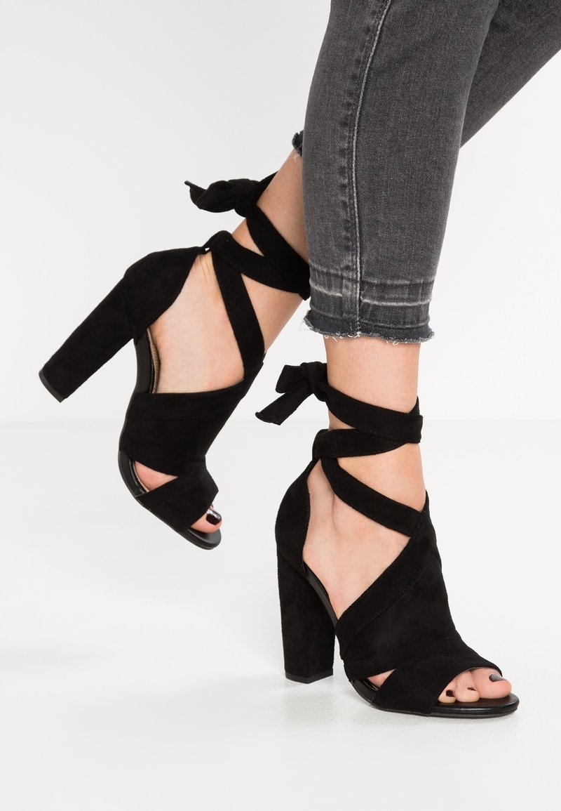 Jambes en jean gris court portant des talons hauts en daim noir avec des talons épais et des sangles autour de la cheville attachées en nœuds.