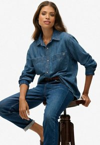Denim overhemd met lange mouwen, knoopsluiting aan de voorkant en twee borstzakken. Draag met bijpassende blauwe denim jeans. Soepele textuur, klassieke pasvorm.
