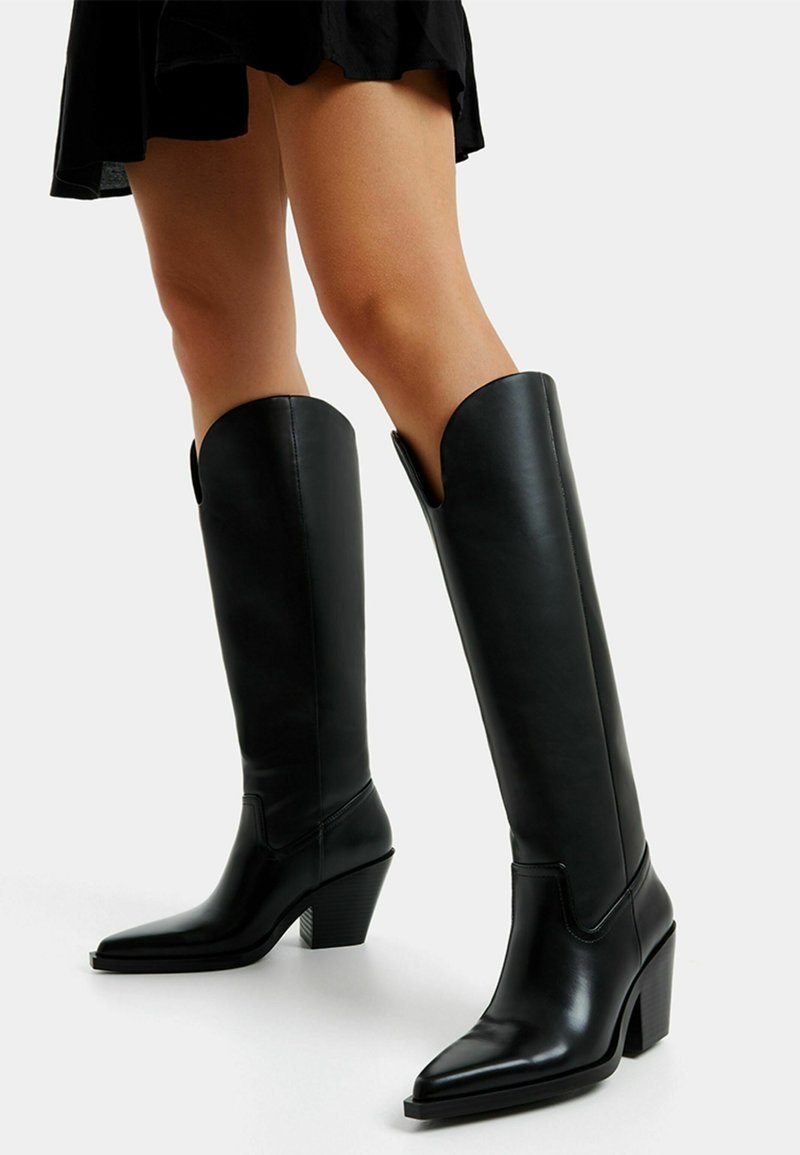 Bottes noires en cuir hautes jusqu'aux genoux avec un bout pointu et un talon carré, présentant une texture lisse et une découpe en V en haut.