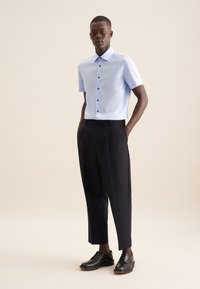 Camicia blu chiara a maniche corte con bottoni scuri, abbinata a pantaloni neri con pieghe e scarpe nere stringate, su uno sfondo neutro.