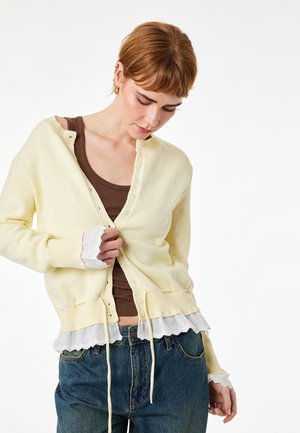 Jong persoon met kort haar die een lichtgeel vestje dichtknopen over een bruin hemdje en een blauwe spijkerbroek, tegen een effen witte achtergrond.