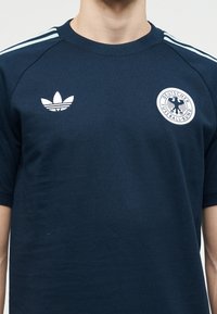 Temno modra športna majica z svetlo modrimi ramenskimi trakovi, Adidas logotip na levi strani prsnega koša in emblem Deutscher Fussball-Bund na desni strani prsnega koša.