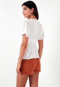 Witte blouse met korte fluttermouwen, gathered taille en een zachte textuur, gecombineerd met roestkleurige shorts, gezien van achteren.