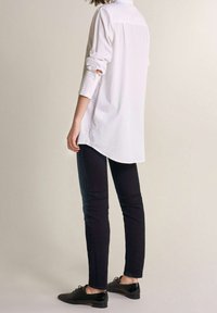 Biała koszula z długim rękawem, z podwijanymi rękawami, o zaokrąglonym dolnym wykończeniu, w połączeniu z ciemnymi dżinsami i czarnymi butami, podkreślająca swobodny, a jednocześnie elegancki wygląd.