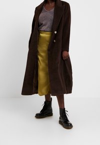 Osoba ubrana w długi ciemnobrązowy płaszcz na błyszczącej szarej bluzce, żółtą satynową spódnicę midi oraz czarne sznurowane botki stojąca na białej podłodze.