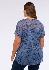 Modrá halenka s vzorem vyrobená z lehké, průsvitné tkaniny. Má krátké srolované rukávy a zakřivený lem, který zdobí jemný vlnkový design.