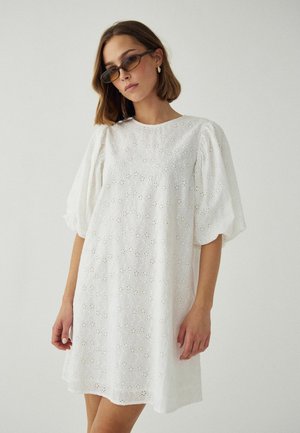 Kobieta w prostokątnych okularach przeciwsłonecznych i białej sukience z bufiastymi rękawami z ażurowej tkaniny, stojąca na tle jednolitego jasnego tła.