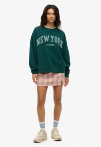 Felpa oversize verde scuro con stampa "NEW YORK", abbinata a una mini gonna a quadri rosa. Sneakers bianche e calzini a righe blu completano il look.