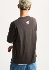 Camiseta de algodón marrón con mangas cortas, que presenta un pequeño diseño gráfico rosa en la parte posterior. Corte holgado con escote redondo.