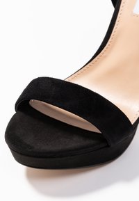 Sandale à talon haut en daim noir avec une large bride unique sur les orteils et une semelle intérieure nude rembourrée.