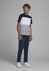 T-shirt in cotone con blocchi di colore blu navy, bianco e grigio, con logo; abbinato a pantaloni jogger blu navy e sneakers bianche. Sfondo semplice.
