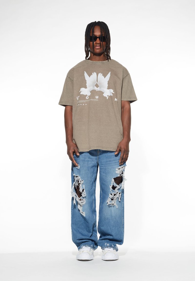 Maslinasta pamučna majica s grafikom bijele golubice i natpisom "YOUTH", u kombinaciji s iznošenim plavim trapericama koje imaju velike poderotine i strgane rubove.