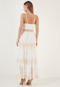 Weißes Maxikleid mit dünnen Trägern, mit einem gestuften Design und beigen Spitzenakzenten. Strukturierter Stoff mit geometrischen Mustern und einem Spitzenbund.