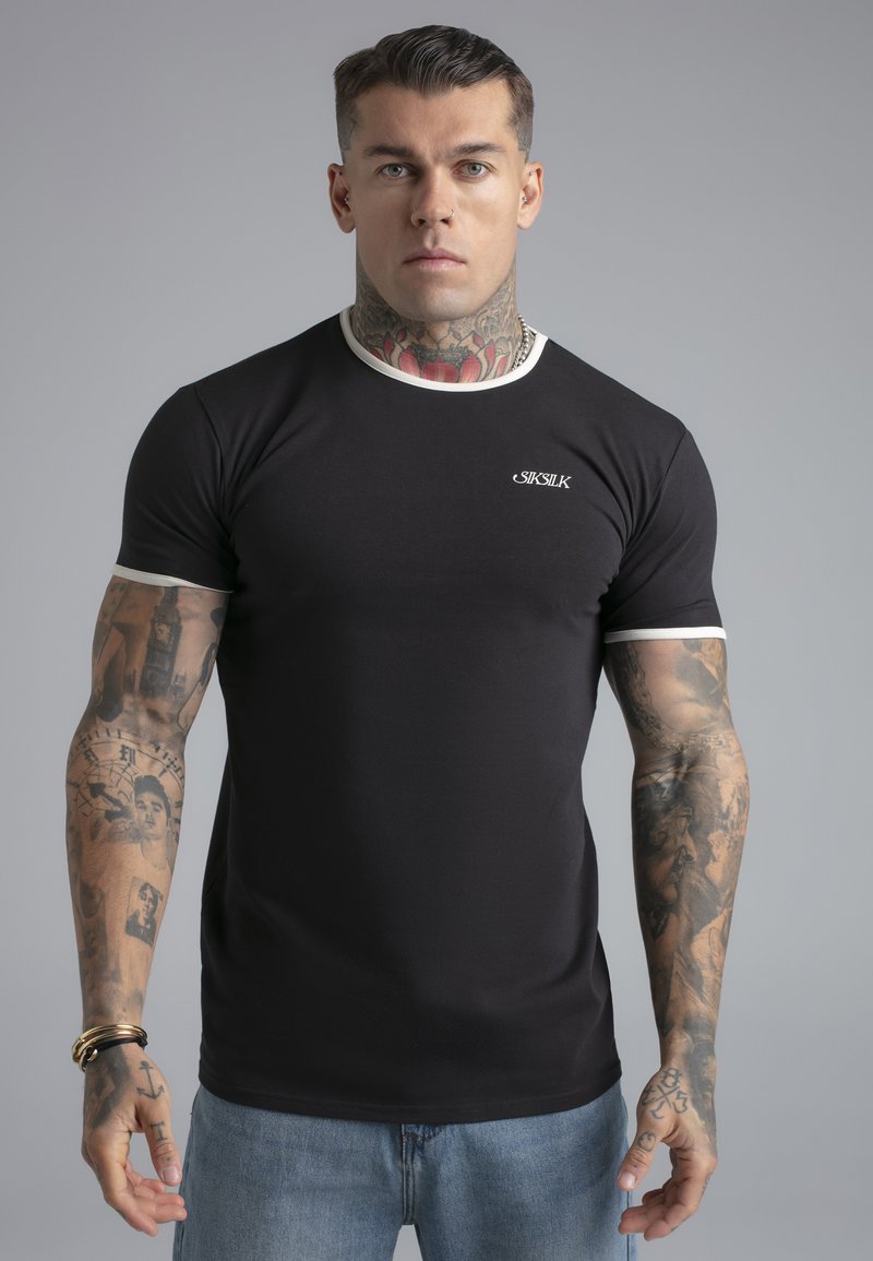 Man met tatoeages op nek en armen die een zwart SikSilk T-shirt en een blauwe spijkerbroek draagt, staand tegen een effen grijze achtergrond.