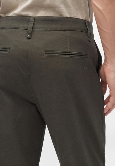 Gros plan d'une personne portant un pantalon vert olive foncé avec deux poches passepoilées à l'arrière et des passants de ceinture, avec une chemise beige rentrée.