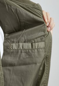 Innenseite einer olivgrünen Jacke mit glatter, glänzender Textur. Verfügt über eine gestickte Tasche und einen Reißverschluss. Eine Hand ist teilweise sichtbar, die den Stoff hält.