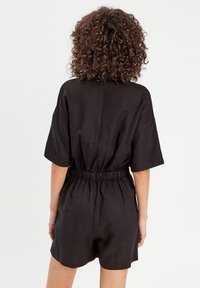 Schwarzer Leinen-Overall mit lässigem Schnitt, kurzen Ärmeln, elastischem Bund und umgeschlagenen Shorts. Glatte Textur und minimalistische Designelemente.