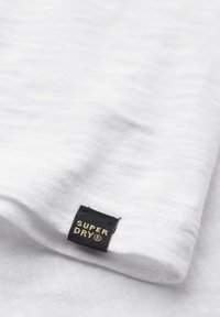 Biała bawełniana koszulka z teksturowaną powierzchnią, z czarną metką wszytą w dół, na której widnieje napis "SUPER DRY" w złotych literach.