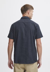 Camisa de manga corta, de color azul oscuro, hecha de un tejido texturizado. Cuenta con un cuello y un dobladillo recto. La vista trasera muestra las costuras de los hombros y un ajuste holgado.