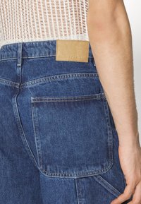 Shorts de mezclilla azul con una superficie texturizada, que presentan una etiqueta de cuero marrón claro en la parte trasera de la cintura y un gran bolsillo trasero en el lado derecho.