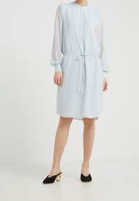 Helles blaues, knielanges Kleid mit vertikalen Streifen, langen durchsichtigen Ärmeln, Rundhals und einer taillierten Taille mit Kordelzug. Schwarze Absatzmules.