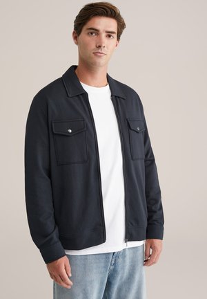 Chaqueta de navy con cremallera y dos bolsillos frontales, fabricada con un material suave. Presenta un cuello alzado y detalles de costura en contraste. Combinada con una camiseta blanca.