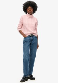 Pull à col roulé rose clair en maille torsadée, jean bleu droit, ceinture noire et chaussures noires à lacets. Le mannequin se tient avec assurance, les mains dans les poches.