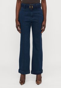 Høyt midjede, vide ben i mørk denim med belte som har en messingspenne, rullede mansjetter og kontrasterende hvite sømmer.