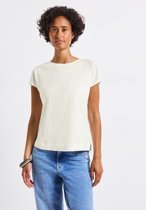 Camiseta de manga corta color marfil con detalles decorativos en los hombros, material de algodón suave, cuello redondo y un corte holgado. Combinada con jeans de color azul claro.