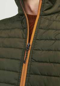 Zielona kamizelka ocieplana z zapięciem na zamek, z pomarańczowymi akcentami wzdłuż zamka. Materiał jest gładki z pikowaną fakturą.