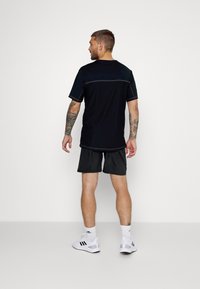Camisa atlética negra de manga corta con costuras en contraste, combinada con pantalones cortos negros. El modelo está de pie de espaldas contra un fondo simple.
