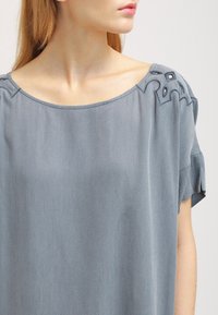 Světle modrý tričko s krátkým rukávem s texturovanou tkaninou, zdobené dekorativním stehem a akcenty na ramenou. Volný střih.