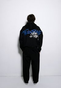 Czarna bluza z kapturem z niebieskim i białym tekstem w stylu graffiti na plecach, w zestawieniu z luźnymi czarnymi spodniami. Wykonana z bawełny, casualowy design.