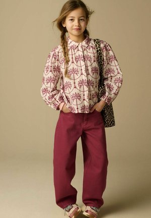 Jeune fille aux cheveux tressés portant un chemisier à motifs, un pantalon bordeaux, des baskets blanches et un sac à bandoulière imprimé léopard, debout les mains dans les poches.