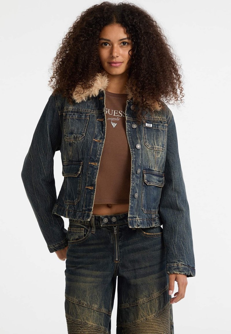 Giacca di jeans con colletto in pelliccia sintetica, lavaggio scuro, design corto, quattro tasche frontali e bottoni in tonalità argentata. Il modello indossa un top marrone e pantaloni di jeans.