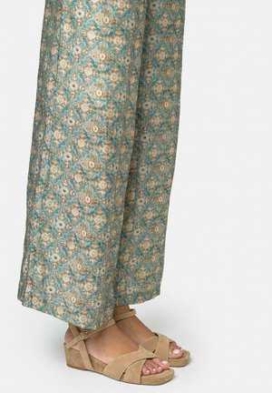 Pies con sandalias cuña beige con tiras en el tobillo y pantalones anchos azules con patrones florales.