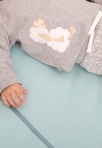 Sweat-shirt gris avec broderie d'un nuage et d'un personnage endormi, en matière coton, avec des poignets élastiques, associé à un pantalon à motif, sur un tapis bleu doux en dessous.