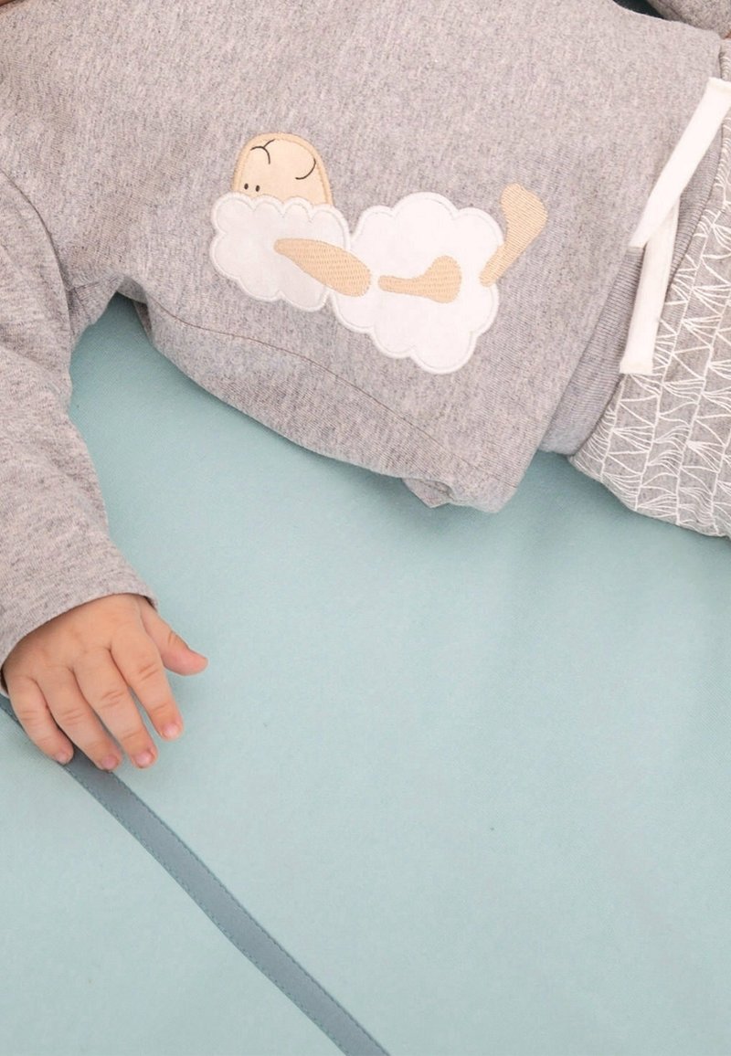 Sweat-shirt gris avec broderie d'un nuage et d'un personnage endormi, en matière coton, avec des poignets élastiques, associé à un pantalon à motif, sur un tapis bleu doux en dessous.