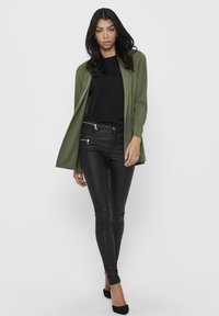 Cardigan aberto verde-oliva sobre uma blusa preta, combinado com calças skinny pretas semelhantes a couro e saltos pretos. Design simples, sem padrões.