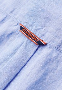 Tejido azul claro con un acabado texturizado, que presenta una etiqueta cosida en azul marino con letras naranjas que dicen "SUPERDRY."