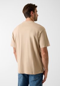 Beżowy bawełniany t-shirt z krótkimi rękawami, luźnym krojem, okrągłym dekoltem i bocznymi rozcięciami zapewniającymi swobodę ruchów. Widok od tyłu.