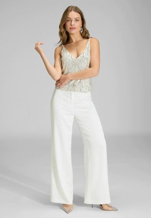 Frau steht in einem ärmellosen, mit Pailletten besetzten Oberteil, weißen Hosen mit weitem Bein und klaren, spitzen Absätzen vor einem schlichten grauen Hintergrund.