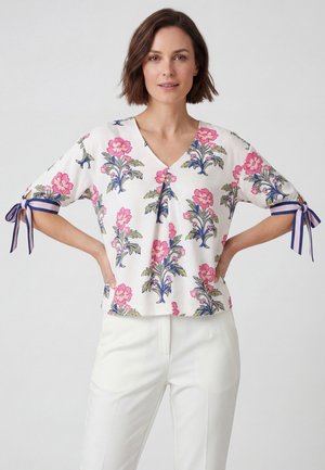 Frau trägt eine weiße Bluse mit rosa Blumendruck und marineblau gestreiften Schleifen an den kurzen Ärmeln, kombiniert mit weißen Hosen.
