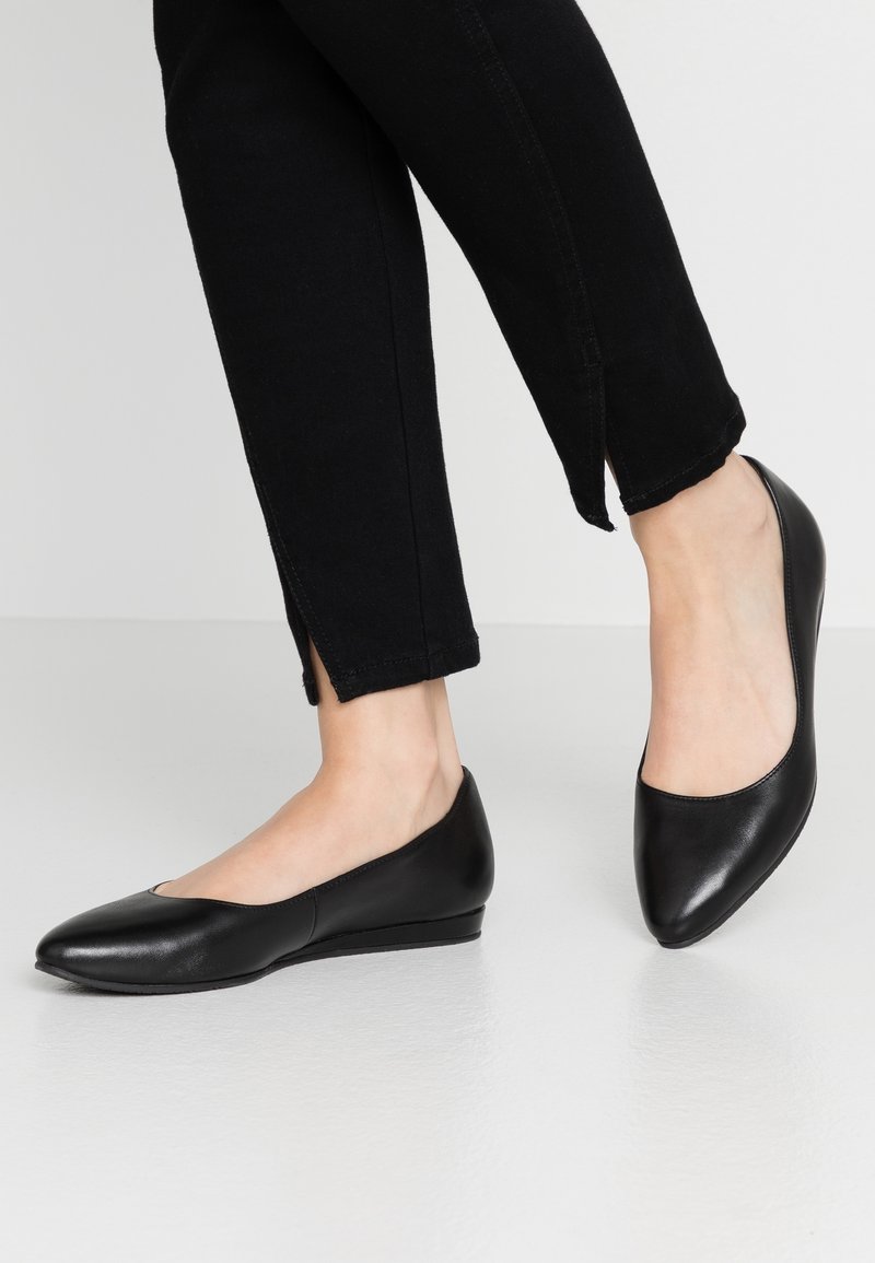 Personne portant des chaussures plates noires à bout pointu associées à un pantalon noir slim comportant de petites fentes latérales aux chevilles.