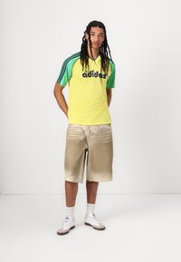 Rumena in zelena Adidas majica z črnim logotipom, v kombinaciji s bež kratkimi hlačami. Model nosi bele superge s sivimi poudarki in bele nogavice.