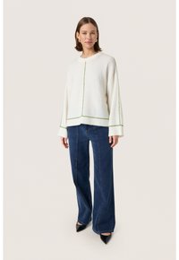 Hvid strikket sweater med grøn kontrastsyning; løs pasform, nedfaldende skuldre. Sammen med mørkeblå denim brede bukser og sorte hæle.