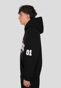 Čierna mikina s kapucňou s veľkou grafikou na prednej strane, rebrovanými manžetami a číslom "01" na rukáve. Mäkká látka a nastaviteľná kapucňa.