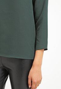 Mörkgrön, lätt top med rund halsringning och trekvartsärmar, kombinerad med glänsande svarta byxor. Slät yta, minimal detaljer.