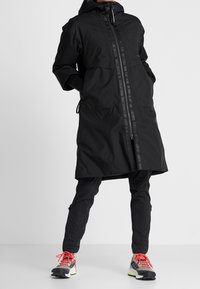 Abrigo largo impermeable negro con cierre de cremallera, capucha ajustable y bolsillos laterales, llevado sobre pantalones negros y zapatillas multicolor.