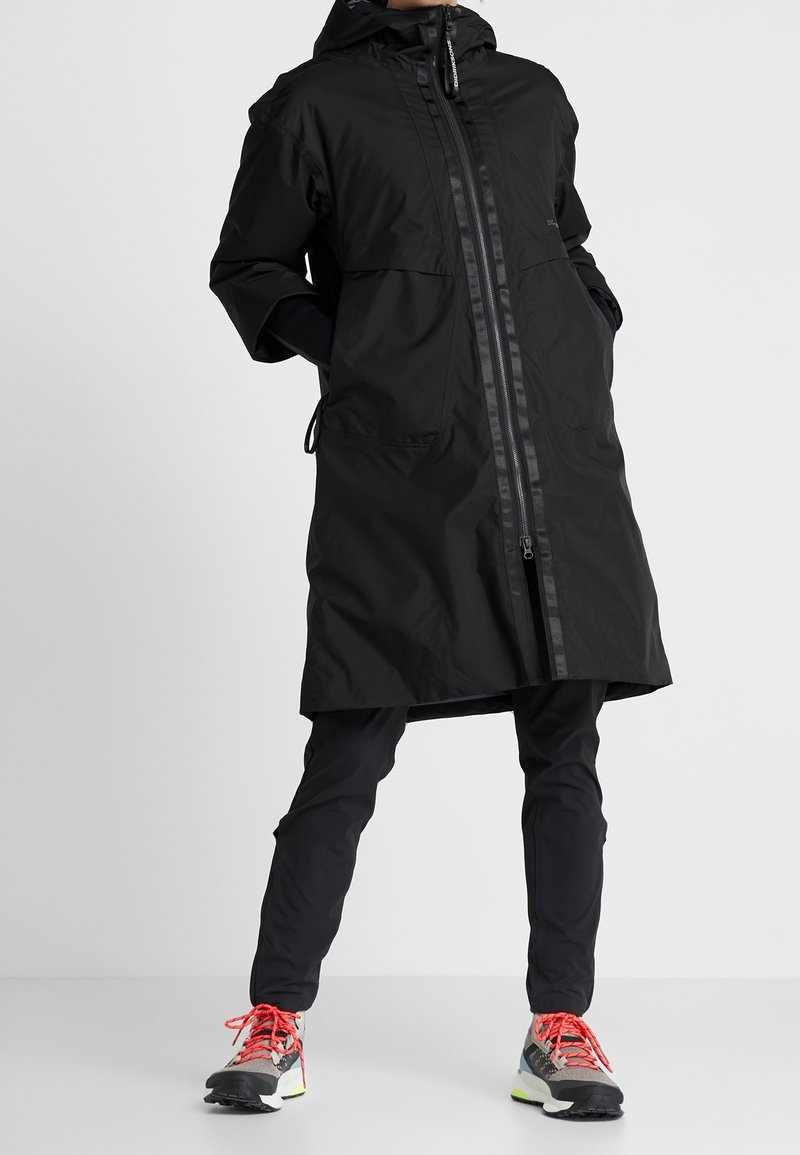 Abrigo largo impermeable negro con cierre de cremallera, capucha ajustable y bolsillos laterales, llevado sobre pantalones negros y zapatillas multicolor.